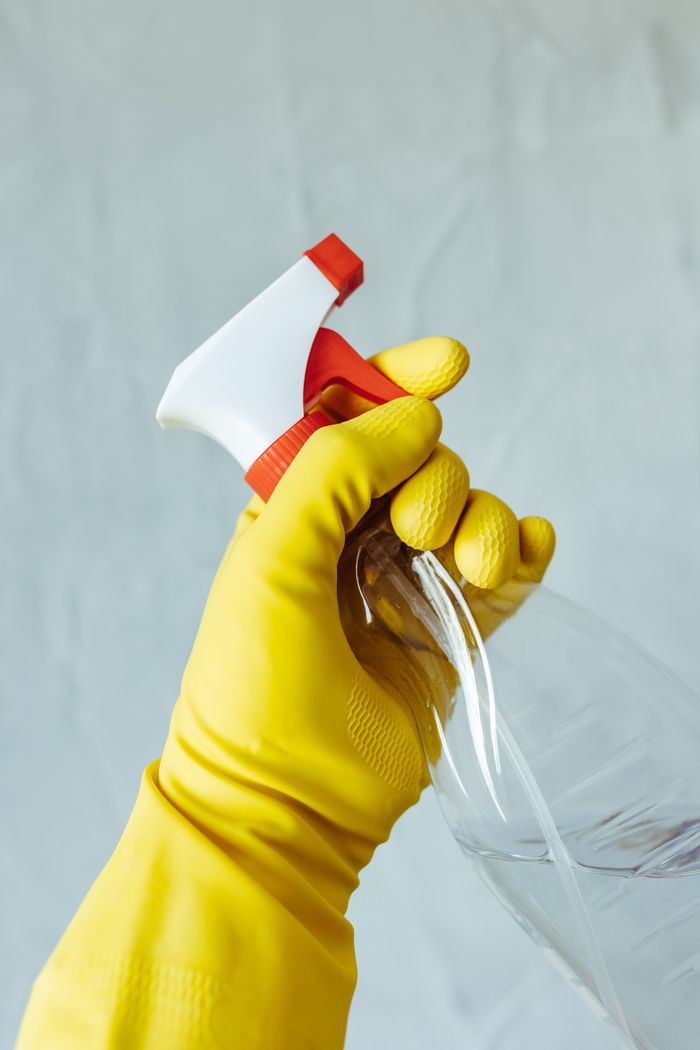 A hand in a yellow rubber glove is holding a red and white spray bottle against a plain background.
